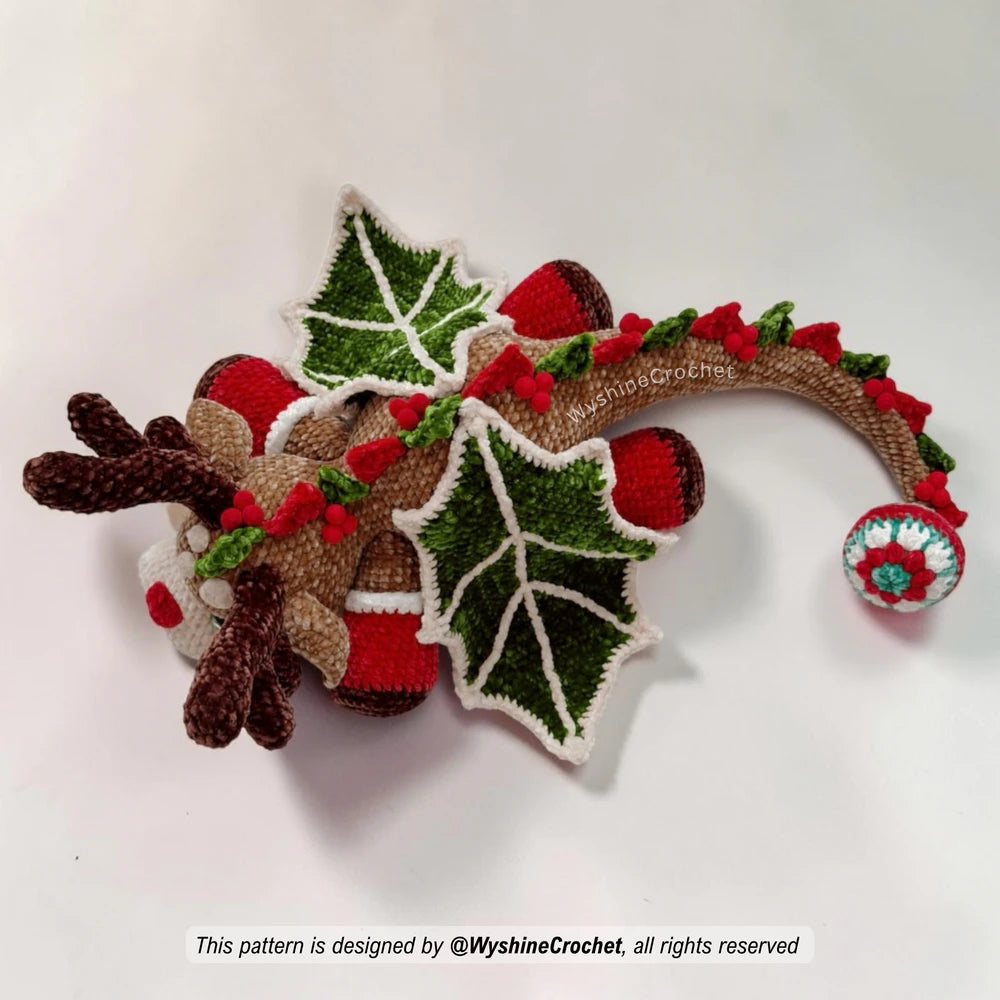 Holiday dragon crochet pattern with reindeer face, red nose, green wings, pom-poms, and cozy chenille yarn texture
